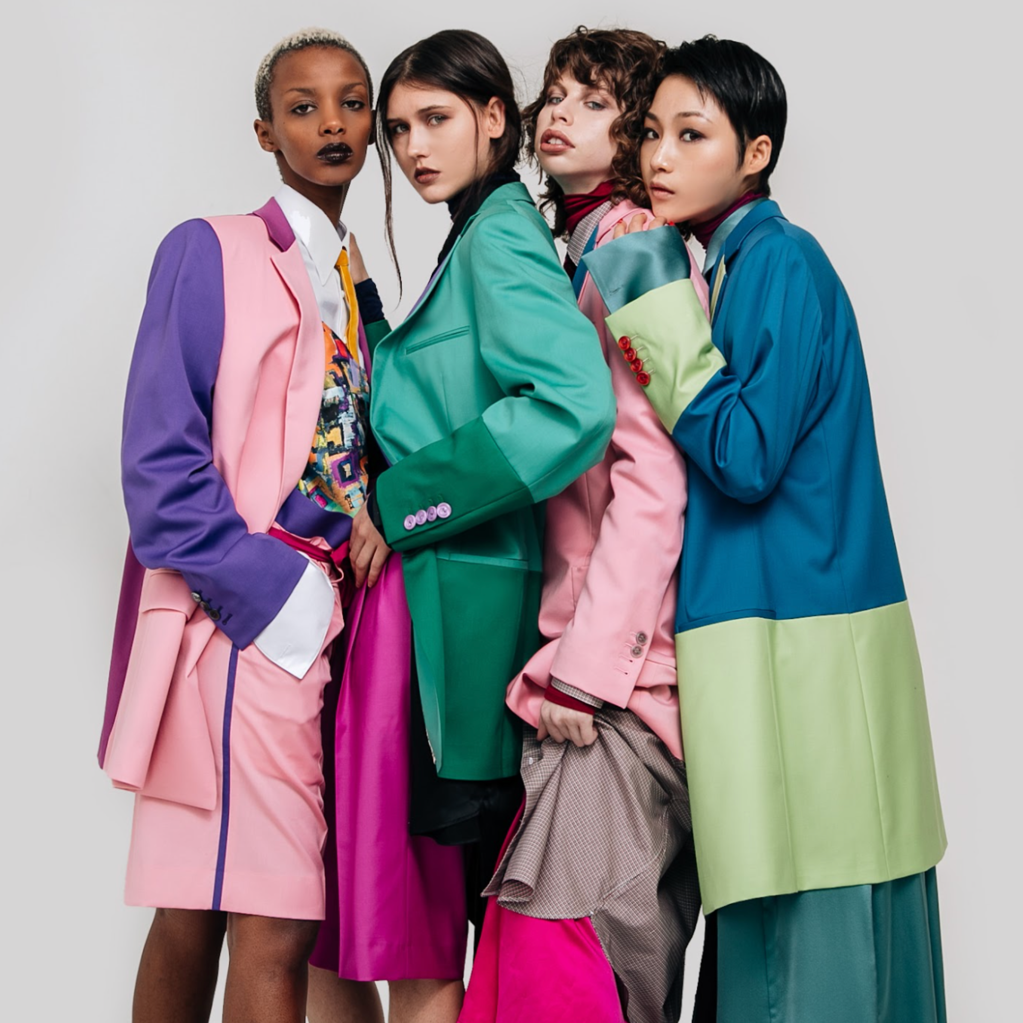 Four models wearing colorblock tailored blazers in pink, purple, green, and blue with contrasting panels, styled with coordinated tailoring pieces, photographed against a light studio background.