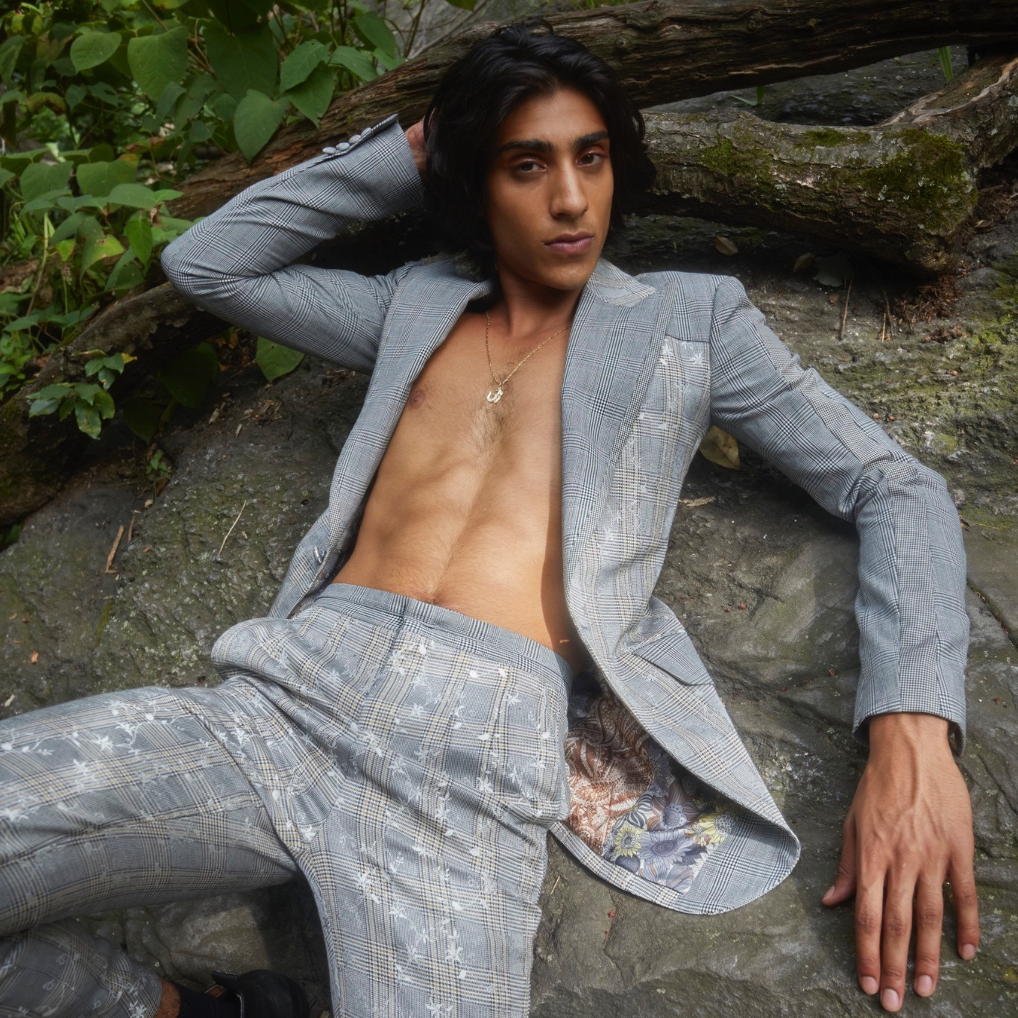 Model wearing a light grey floral plaid tailored blazer with deep V neckline, paired with matching trousers, photographed outdoors reclining on stone and tree roots in a wooded setting.
