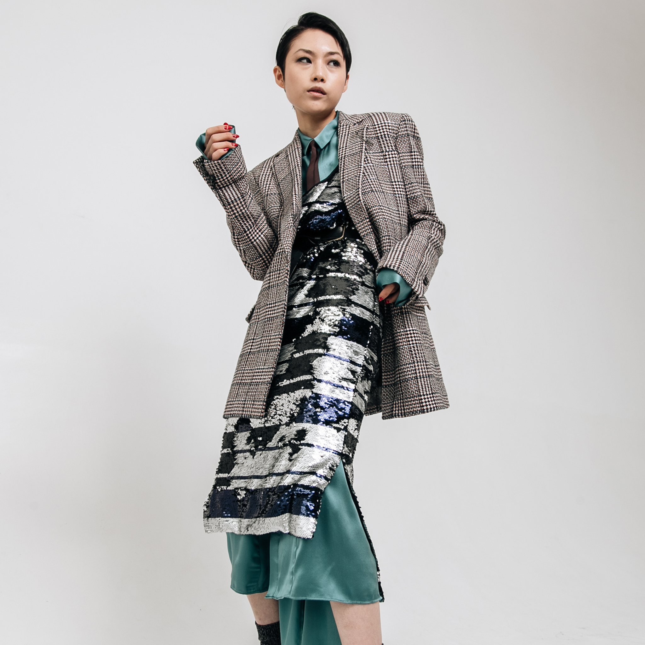 Model wearing a brown and black plaid tailored blazer layered over a silver and navy sequin dress with teal satin underlayer, photographed against a white studio background.