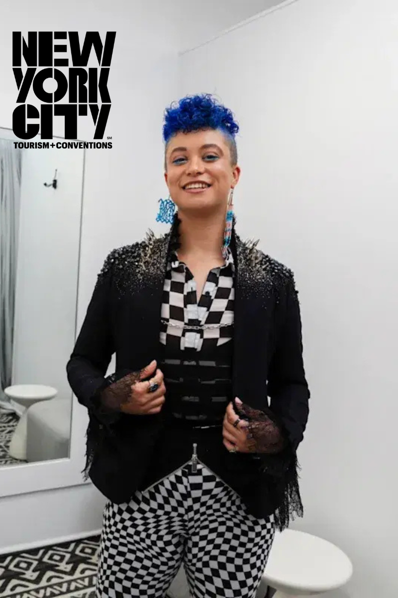A person with bright blue hair stands in a modern white fitting room, wearing a black blazer and bold black-and-white checkered clothes, smiling confidently toward the camera.