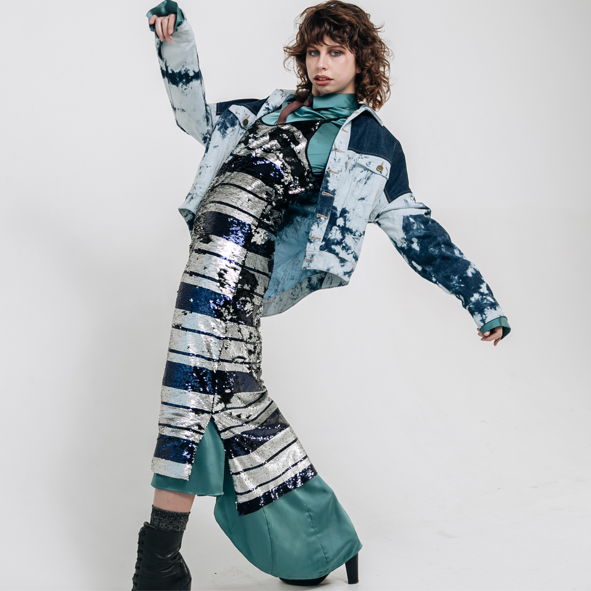 Model wearing an indigo patchwork denim jacket layered over a striped silver and navy sequin midi dress with teal satin underlayer, photographed against a white studio background.